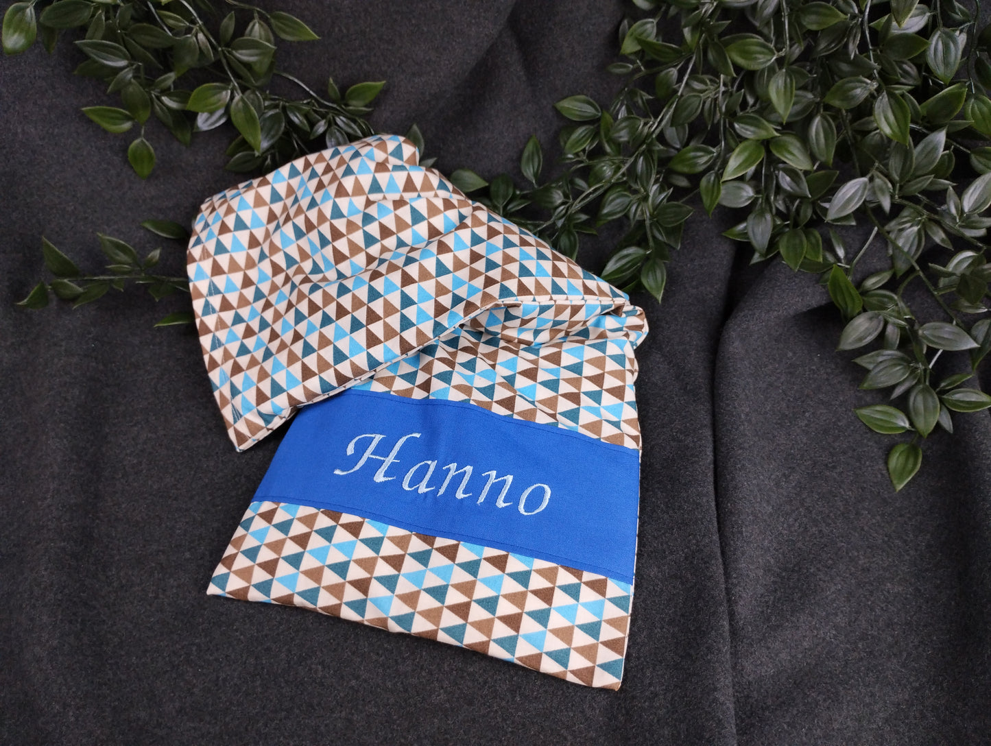 personalisiertes Körnerkissen mit unserem Dreiecksmotiv in blau braun. Darauf abgebildet der gestickte Name Hanno in hellblau auf mittelblauem Stoff